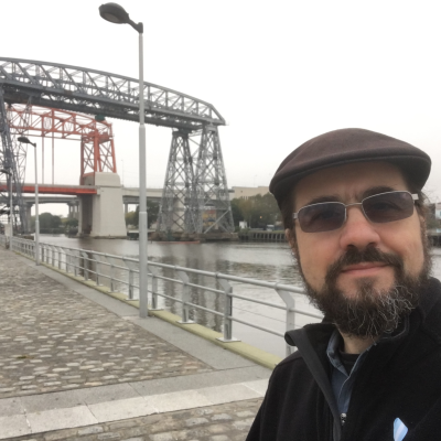 a man with a beard and a cap stands in front of industrial bridgework spanning a canal